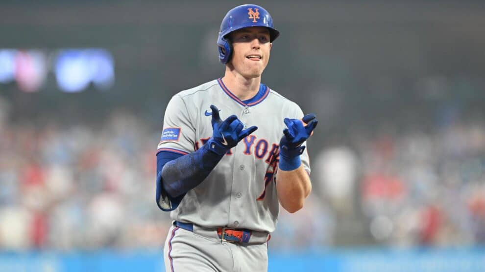 New York Mets player in gray away uniform and blue helmet, gesturing with blue gloves on the field.